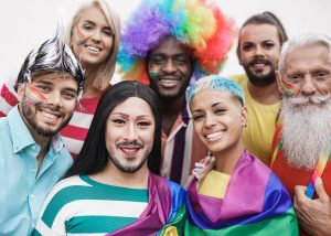 Group of seven people, all ages, genders, sexualities, and nationalities dressed up in colourful gay pride festival attire.