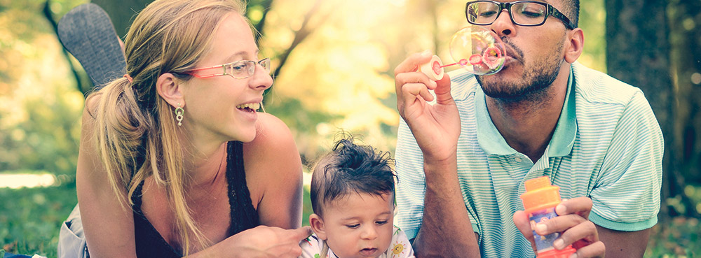 Young couple in their late twenties laying on a picnic blanket blowing bubbles with their young child